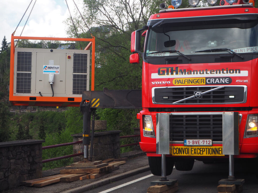 Rode kraanwagen hijst de rooftop airco-unit in oranje frame op zijn plaats in Houffalize