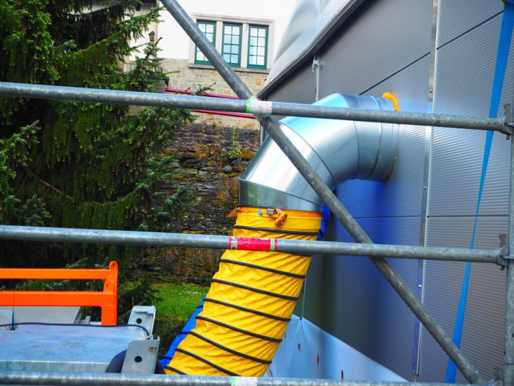 Gele flexibele luchtslang op een aluminium bocht aan de gevel, achter een stelling