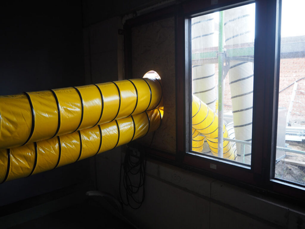 Twee gele luchtslangen die via een raamopening warme lucht binnenbrengen in het studentengebouw